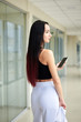 © Natalia - Young brunette woman, wearing white pants and black top, holding jacket in hand, looking at cell phone in light passageway of office building. Businesswoman on lunch break. Corporate culture concept.