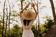Brunette woman in a forest, wearing a summer dress and a straw hat, dancing in the sun.