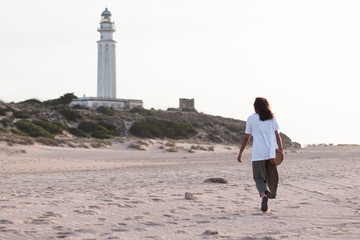 Young brown woman walking on the beach on a sunset to a lighthouse.