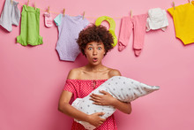Stunned Curly Haired Mother Holds Infant Baby, Finds Out About Childs Serious Illness, Keeps Mouth Widely Opened, Poses Against Washed Clothes On Rope, Pink Background. Newborn, Moterhood Concept