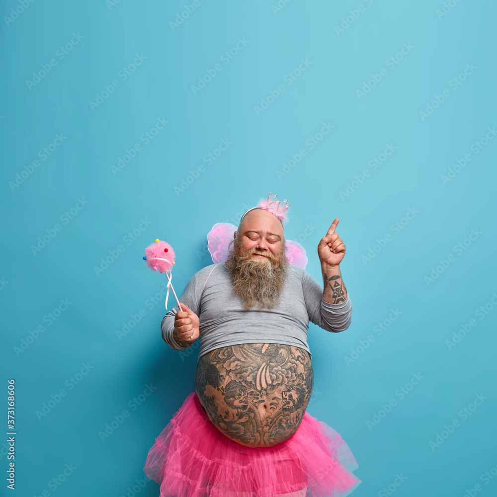 People, holiday, fun concept. Joyful obese man wears princess outfit, poses with magic wand, moves actively, has big tattooed stomach stands closed eyes points above on copy space blue background - obrazy, fototapety, plakaty 