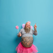 © WHstudio Leushin N - People, holiday, fun concept. Joyful obese man wears princess outfit, poses with magic wand, moves actively, has big tattooed stomach stands closed eyes points above on copy space blue background