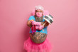 Impressed bearded man maid, crosses hands over big tattooed belly, holds detergent and plunger for cleaning, dressed in funny fairy costume, busy with household duties, isolated on pink background