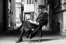Stylish Long-haired Girl In A Long Black Coat, Cap And Green Dress Is Reading A Book In A Shabby Courtyard...Typical Petersburg Courtyard