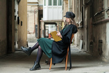 Stylish Long-haired Girl In A Long Black Coat, Cap And Green Dress Is Reading A Book In A Shabby Courtyard...Typical Petersburg Courtyard
