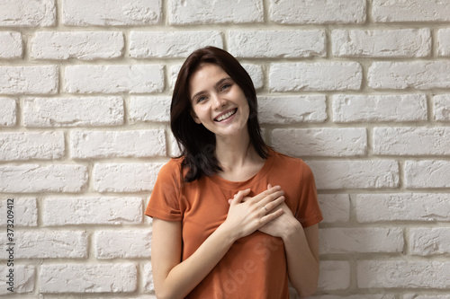 Portrait of smiling young Caucasian woman isolated on white brick wall background hold hands at heart chest show gratitude support, happy millennial girl volunteer feel thankful demonstrate care
