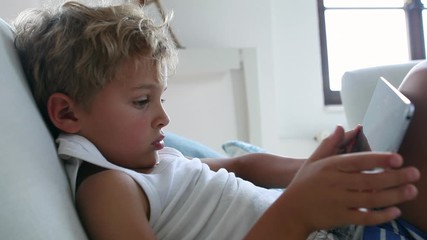 Wall Mural - Child sitting in sofa looking at tablet device. Young boy using tech tablet