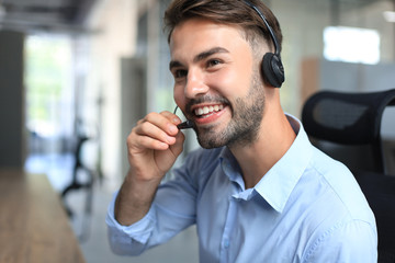 Cheerful young support phone male operator in headset, at workplace while using laptop, help service and client consulting call center concept.