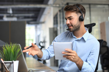 Sticker - Smiling male call-center operator with headphones sitting at modern office, consulting online information in a laptop, looking up information in a file in order to be of assistance to the client.