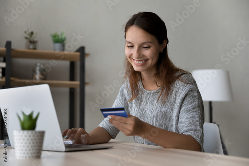 Smiling millennial Caucasian female sit at table at home make online payment purchase using credit card, happy young woman shopping on internet, pay on web with secure banking service system on laptop