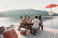 Group Of Friends Sitting At The Table And Have Dinner On A Pier With Beautiful Landscape Of The Lake