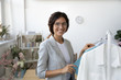 © fizkes - Portrait of smiling pleasant beautiful young female fashion designer in eyeglasses standing near new clothing collection hanging on rack in modern atelier showroom, creative profession dressmaking.