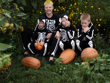 Friendly Family In Carnival Costumes Of Skeletons Sit Among The Thickets Of Sunflower. Halloween Eve. Mom And Three Boys With Pumpkins