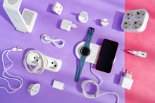 The Concept Of Wireless Charging Of Mobile Gadgets. The Smartphone And Smartwatch Are On The Wireless Charging Pad. There Are Various Adapters, Cables And Chargers Nearby. Colored Paper Background.