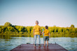© Georgii - Cool kids brothers holding hands look at the river staying on dock. Back view