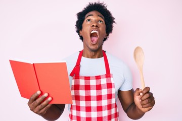 Handsome african american man with afro hair wearing professional baker apron reading cooking recipe book angry and mad screaming frustrated and furious, shouting with anger looking up.