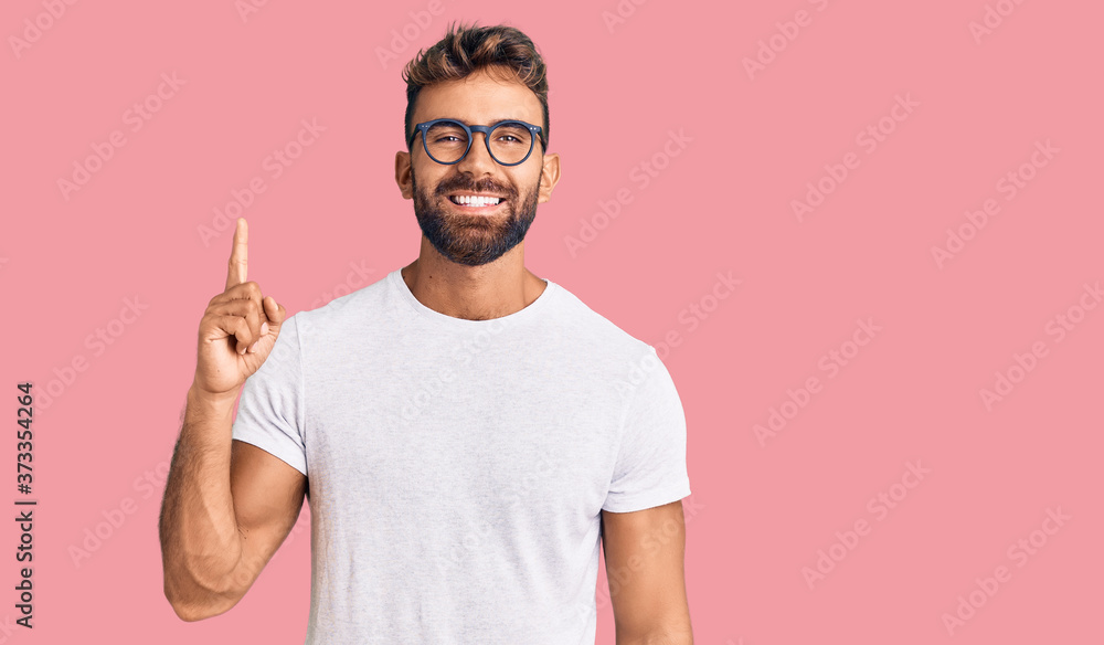 Young hispanic man wearing casual clothes and glasses showing and pointing up with finger number one while smiling confident and happy. - obrazy, fototapety, plakaty 