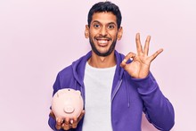 Young Latin Man Holding Piggy Bank Doing Ok Sign With Fingers, Smiling Friendly Gesturing Excellent Symbol