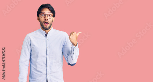 Fotografía Handsome hispanic man wearing business shirt and glasses surprised pointing with hand finger to the side, open mouth amazed expression
