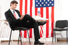 US Politics. American Politician Is Sitting Next To Flag. Politician Writes Something. USA Flag In Campaign Headquarters Of Politician. Concept - Career In US Politics. United States Of America