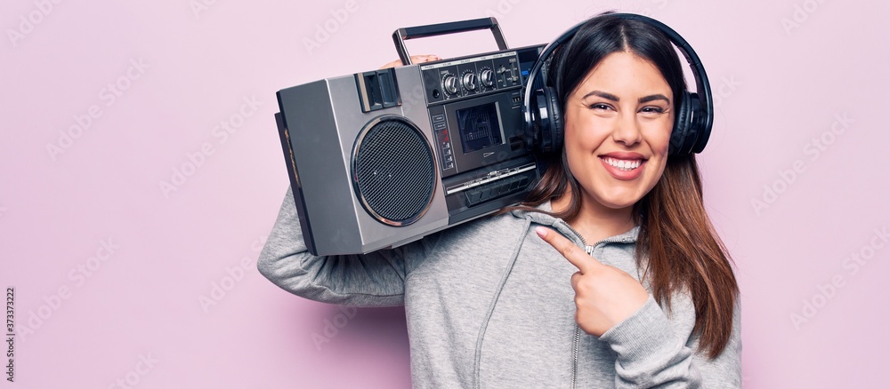 Young beautiful brunette woman listening to music using vintage boombox and headphones smiling happy pointing with hand and finger - obrazy, fototapety, plakaty 
