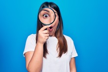 Young Beautiful Brunette Woman Using Magnifying Glass Over Isolated Blue Background Scared And Amazed With Open Mouth For Surprise, Disbelief Face