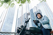 © DragonImages - Handsome serious Vietnamese young man in helmet sitting on motorcycle with smartphone in hands, skyscrapers in background
