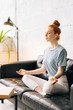 © dikushin - Loving redhead young woman meditating and relaxing in lotus pose while sitting on soft couch. Business lady taking break from work at computer and documents