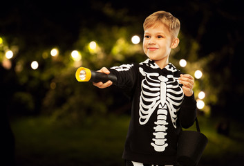 Poster - halloween, holiday and childhood concept - happy boy in black costume of skeleton with bucket of candies and flashlight trick-or-treating over night park and garland lights on background