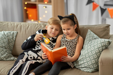 halloween, holiday and childhood concept - little kids in costumes with flashlight reading book at home