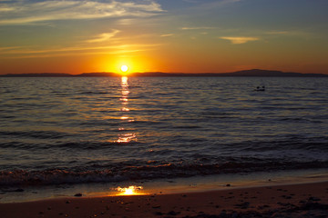 Sunset on Lake Baikal in dark moody colors, sun sets over the horizon above calm waves and 2 gulls on stone in the water