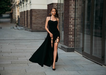 Young Woman In Classic Evening Black Dress. Fashion Photo. Luxury Girl With Long Bare Leg. Glamour Stylish Model. Beautiful Retro Lady Walks On Old City Street. Elegant Makeup Long Wavy Hair Sexy Face