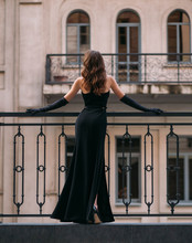 Mysterious Silhouette Of An Elegant Retro Woman. A Lady In Long Black Evening Dress Stands On The Balcony. Rear View, Sexy Back. Long Vintage Gloves. Glamor Hairstyle Hollywood Wave, Light Brown Hair