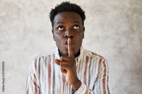 Do not tell anybody. Mysterious young African male looking up, holding fore finger on his lips, saying shh, asking to be quiet, keep silent, sharing secret with you. Consipracy and secrecy concept