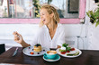 Cheerful caucasian female sitting at cafe interior on breakfast meal laughing eating tasty salads and pancakes, happy hipster girl satisfied with delicious healthy diet nutrition keeping lifestyle