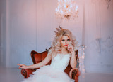 Angry young beautiful girl princess misses sadness loneliness. Queen woman blonde woman long curls hair. Hairstyle with vintage royal crown. Backdrop white room, candelabra romantic lit bright sparks