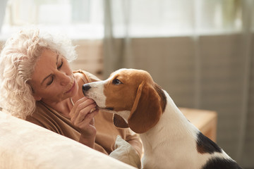 Portrait of white haired senior woman playing with dog and giving him treats while sitting on couch in cozy home interior, copy space