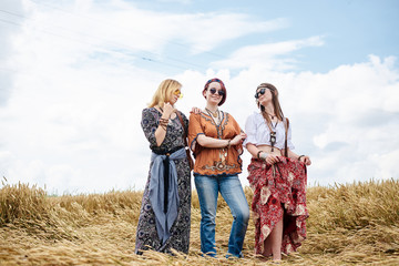 Three hippie women, wearing boho style clothes, standing in the wheat field, smiling, Female friends, traveling together in countryside. Eco tourism concept. Summer leisure time