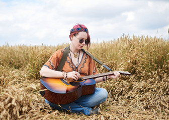 Young hippie women with red hair, wearing boho style clothes, sitting in the middle of wheat field, playing guitar. Travel musician in countryside. Eco tourism concept. Summer leisure activity.