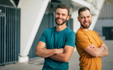 Two happy friends or brothers in colorful t-shirts are standing back to back with crossed arms outdoors