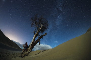 Poster - Male under a dry tree in a sandy area under the night sky with the milky way