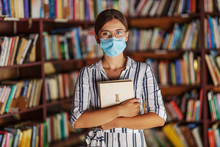 Portrait Of Young Attractive College Girl Standing In Library With Face Mask On Holding A Book. Studying During Covid 19 Concept.