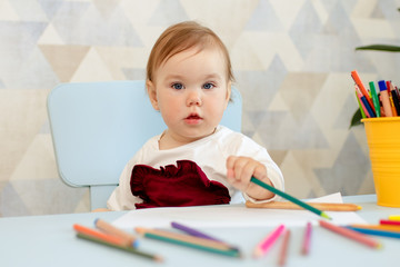 Happy little one year old girl draws with pencils at the table.