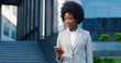 Beautiful young African American woman standing outdoor and texting message on mobile phone at business center. Female tapping and scrolling on smartphone at street. Businesswoman using telephone.