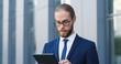 © VAKSMANV - Handsome Caucasian young businessman in glasses, suit and tie using tablet device at street. Man in business style tapping and scrolling on gadget computer. Outdoor. Close up.