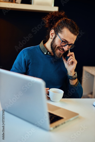 Young smiling handsome bearded hipster with curly hair sitting in his home office, having phone call and drinking coffee during corona virus.