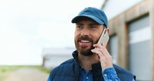 Close Up Of Handsome Cheerful Caucasian Man With Beard And In Hat Standing Outdoor And Talking On Mobile Phone At Wooden Shed. Happy Male Farmer Speaking On Cellphone. Telephone Call. Conversation.