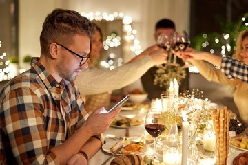 Wall Mural - holidays, technology and people concept - young man in glasses with smartphone at christmas dinner party with friends at home