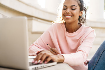 Wall Mural - Image of cheerful african american student girl doing homework with laptop