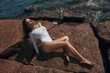 © Dmitry Tsvetkov - High fashion oytdoor portrait. Sexy tanned caucasian woman with long brunette straight hair in white body and sunglasses sitting at huge stone or rock at seacoast on a sunny day